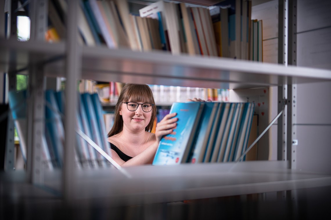 Schülerin in Bibliothek blickt durch Regal mit Büchern